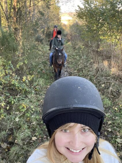 Rebecca smiles at the camera wearing a black hat and protective helmet; behind her, her friends ride two brown horses. 