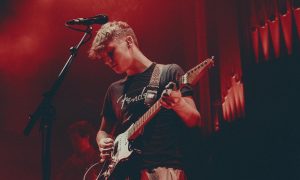 Ben Walker stands on stage with a Fender guitar and microphone against a red backdrop.