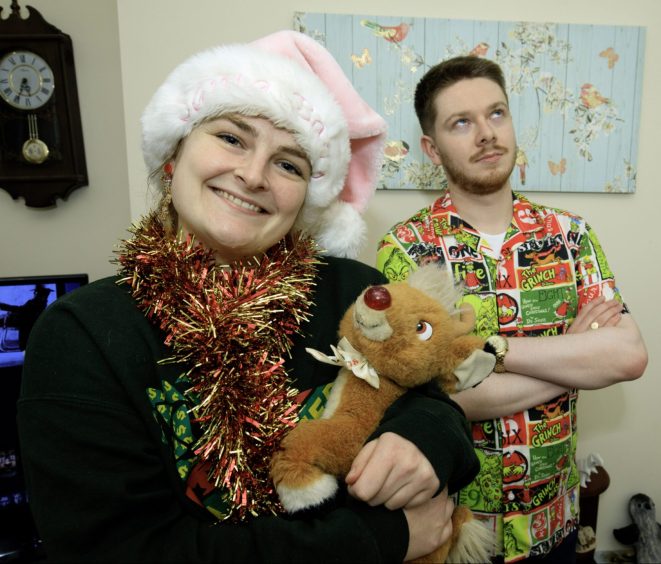 Becky Fairly smiles at the camera, sporting a pink Santa hate, red and gold tinsel scarf and a cuddle Rudolph plush toy, as her flatmate John stands in the background rolling his eyes, wearing a Grinch-themed button-down shirt. 