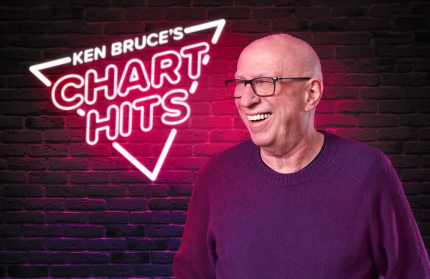 Ken Bruce smiles in front of neon sign that reads 'Ken Bruce's Chart Hits'