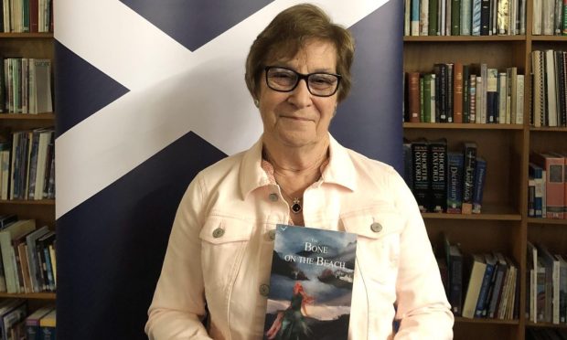 Image shows author Fiona Gillan Kerr holding a copy of her novel The Bone on the Beach. She is standing in front of a Scottish flag, is wearing glasses and smiling.