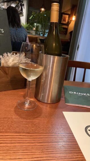 An example of the food and drink on offer in Broughty Ferry. Image shows a glass of white wine with the bottle behind it, with a drinks menu set on the table. 