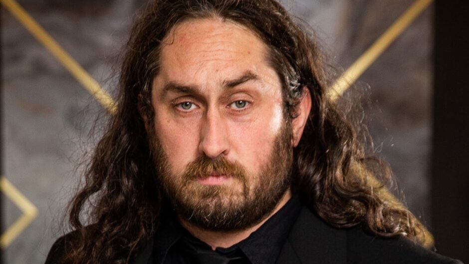 Ross Noble (a white man with long dark hair and a beard) stares directly into the camera in front of a dark grey background with a golden X-shaped pattern. 