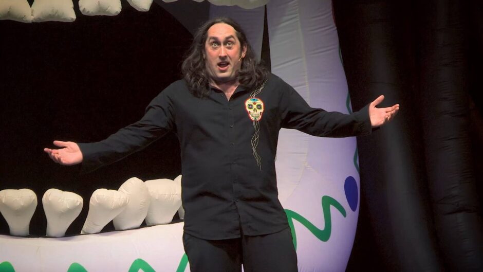Ross Noble stands with his arms out in front of the mouth of a giant inflatable skull. 