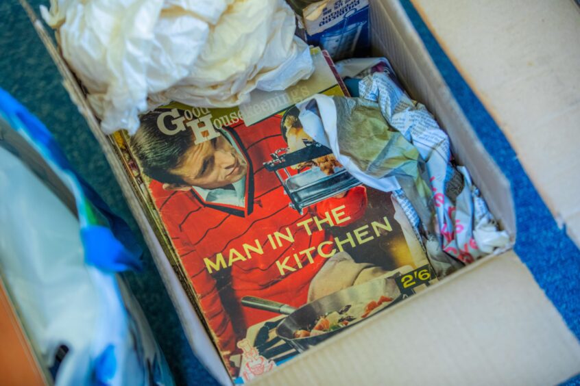 Box containing books and magazines, on the top is a 50s style Good Housekeeping publication called Man in the Kitchen