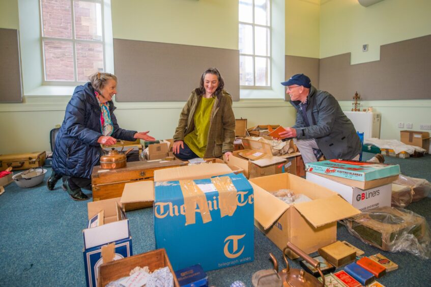 Three people kneeling on floor of Crieff Town Hall surrounded by packing boxes and objects