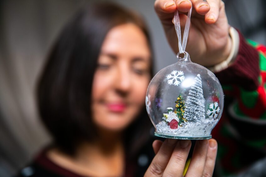 Caris (blurred in the background) holds a delicate glass Christmas bauble with a tiny tree and cottage inside up to the camera. 