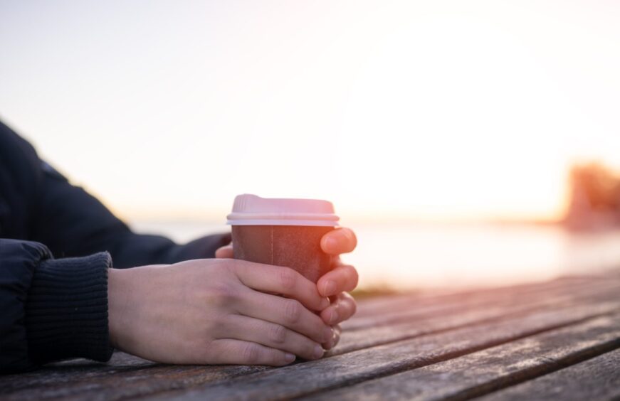 person holding takeaway coffee cup on outside picnic table