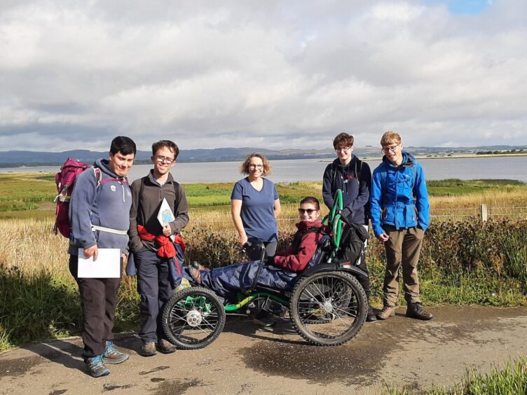 The five group members in outdoor gear in the Scottish countryside with Kayleigh in an all-terrain wheelchair.