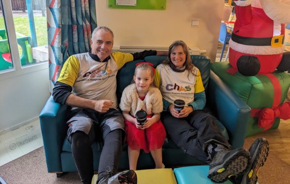 Bob and Deborah Gulliver in Rachel House, Kinross, with a little girl. The three are sitting on comfy chairs with a blow up Santa and colourful decorations all around them.