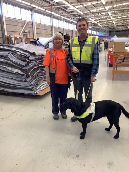 Woman and man in high-vis vests in a warehouse environment. The man has a black Labrador guide dog.