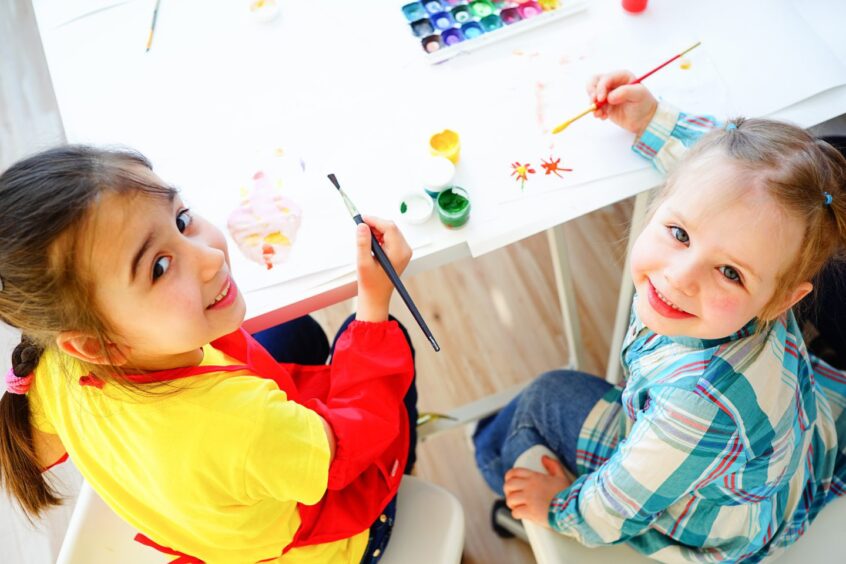 Children sitting at a table with paints. They are looking up and smiling.
