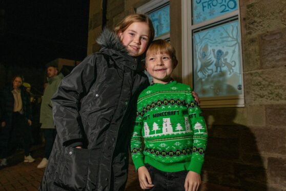 Two young children in warm coat and Christmas jumper outside an illuminated window with a Jack Frost design in the Craigie area of Perth.