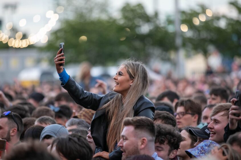 A girl sitting on someone's shoulders in a concert crowd takes a selfie