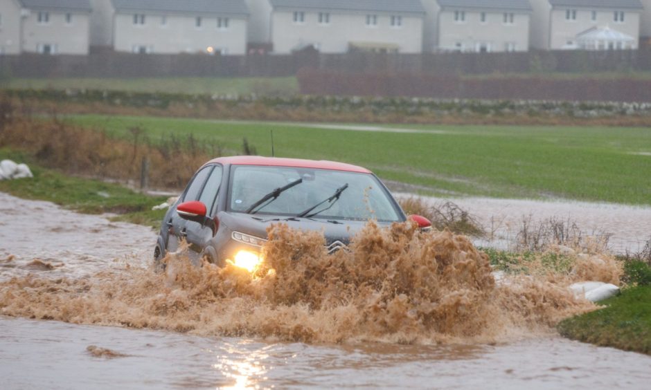 Car driving through puddle with flood water splashing over bonnet and pouring into neighbouring field on outskirts of Perth
