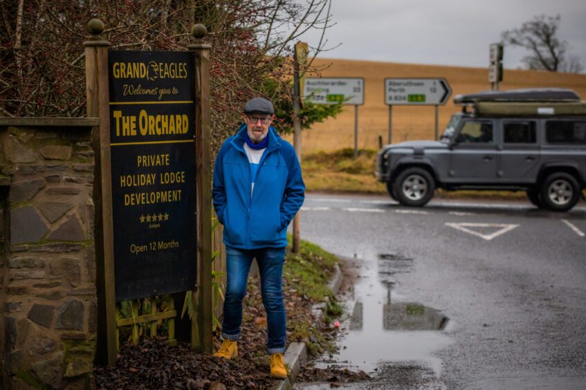 Alasdair Gow standing next to entrance sign for The Orchard private holiday lodge development on the Shinafoot road with a LandRover passing behind him.