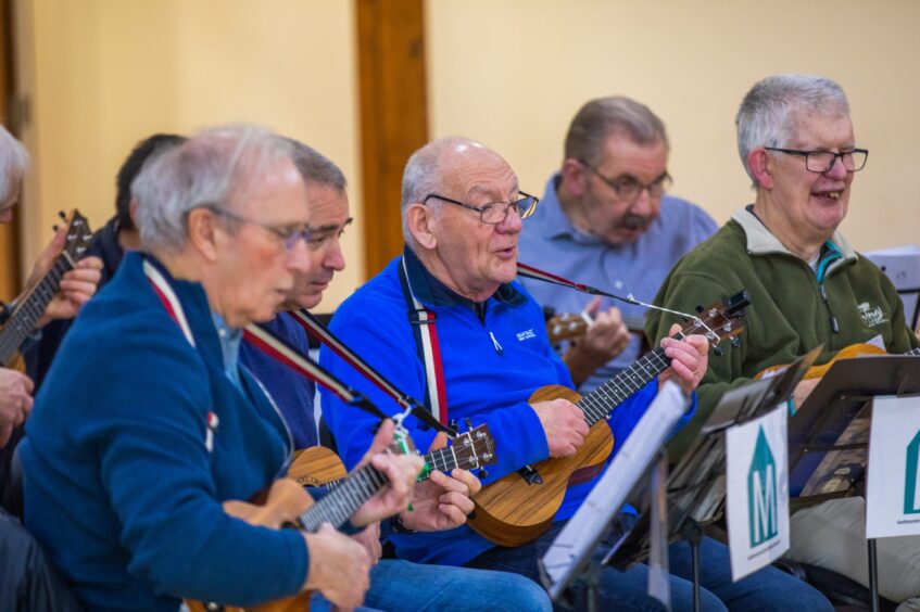 Row of elderly men playing ukuleles and singing with sheet music in front of them.