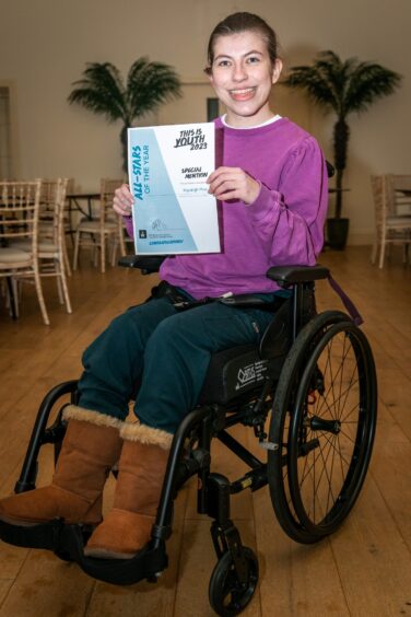 Kayleigh Ptak in wheelchair, holding certificate with big smile