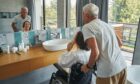 Senior lady in wheelchair touching her face in front of bathroom mirror while elderly man stands behind her