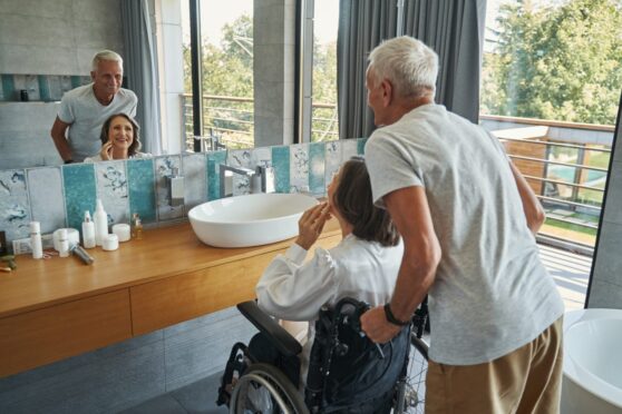 Senior lady in wheelchair touching her face in front of bathroom mirror while elderly man stands behind her