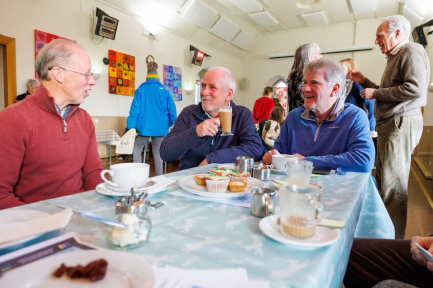 Douglas Fraser holding a frothy coffee at a table with two other men, with a large number of people seated and standing behind them in Glenfarg community centre.
