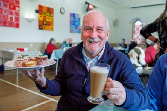 Douglas Fraser, with white hair and beard. smiling and holding a coffee and cake to the camera at the weekly coffee lounge in Glenfarg community centre.