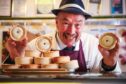 Alan Pirie in butcher gear smiling as he holds two pies to the camera behind the counter in his Newtyle shop
