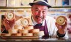Alan Pirie in butcher gear smiling as he holds two pies to the camera behind the counter in his Newtyle shop
