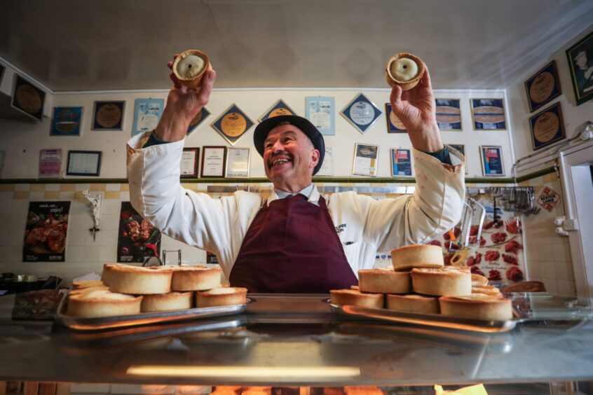 Alan Pirie smiling as he holds two pies up to the camera inside his butcher shop