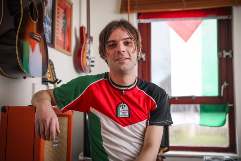 Musician Nick Shane wearing a T-shirt sporting the Palestinian flag, with another Palestinian flag draped over his window in the background to show support for those affected by the war. 