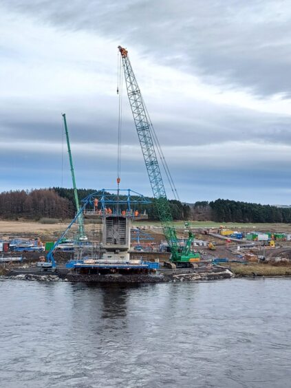 One side of new bridge beside River Tay, with workmen on top, a crane, and associated construction site.
