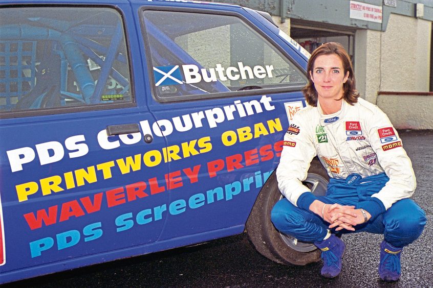 Image shows Jillian Butcher (now Jillian Sheddon) in her motorracing gear. Jillian is crouching in front of her car, which is covered in sponsorship messages.