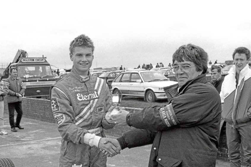 Image shows a black and white photograph of a very young David Coulthard being presented with a trophy after a race at Knockhill Racing Centre.
