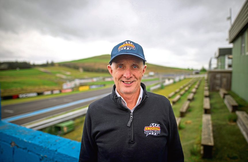 Image shows Stuart Gray, director of events at Knockhill racing track. Stuart is wearing a Knockhill cap and sweater and in standing in the middle of the picture with a view of the race track in the background.