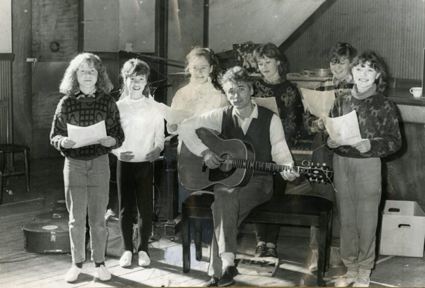 Michael Marra holds a guitar while children holding sheets of paper stand around him