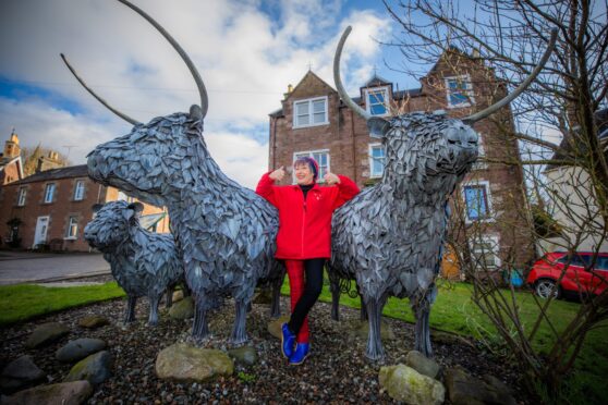 June McEwan giving two thumbs up next to metal Highland sculptures depicting a Highland cow, bull and calf in Crieff.