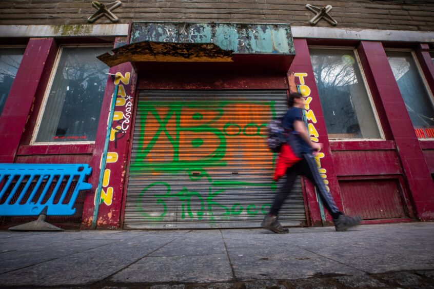 Shows red façade of vacant Toymaster unit on Commercial Street in Dundee. A person is walking its shutters covered in graffiti.