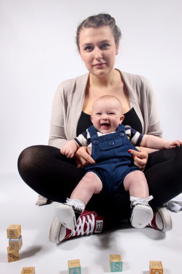 Image shows young mum Amy Deans with her son Mason at Bringing Up Baby playgroup. Amy is sitting cross-legged with her smiley baby on her knees.