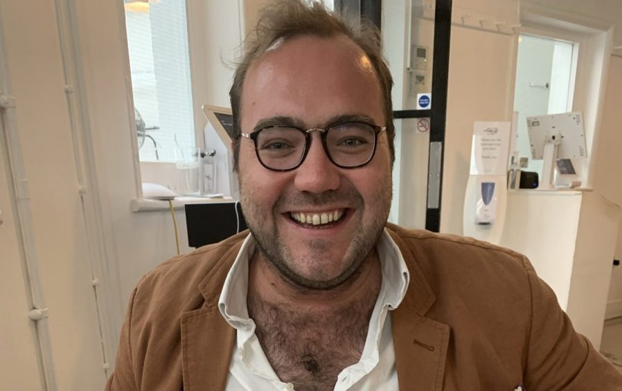 Image shows: columnist and writer Andrew Liddle. He is in his mid 30s, weaing round rimmed glasses, white shirt open at the neck and a tan-coloured jacket. He is smiling broadly.