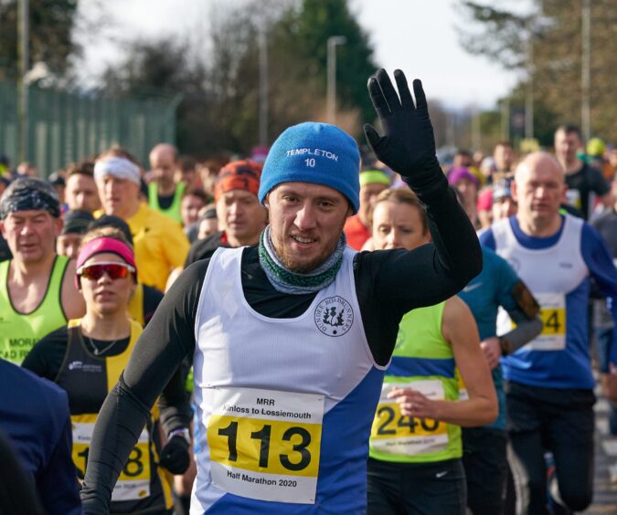 A member of the Dundee Road Runners club waves to the camera as hetakes part in the Kinloss to Lossiemouth Half Marathon, with other runners in the background. 