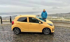 Jack McKewon beside a yellow Kia Picanto at the top of a Dundee hill, with the River Tay in the background.