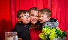 Image shows Amy Deans of Front Lounge Dundee with her sons Mason and Parker. Amy is sitting with her arms around both boys. All three are happy and smiling and Mason is wearing glasses. Amy is wearing a red top and they are in front of a red background.