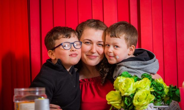 Image shows Amy Deans of Front Lounge Dundee with her sons Mason and Parker. Amy is sitting with her arms around both boys. All three are happy and smiling and Mason is wearing glasses. Amy is wearing a red top and they are in front of a red background.