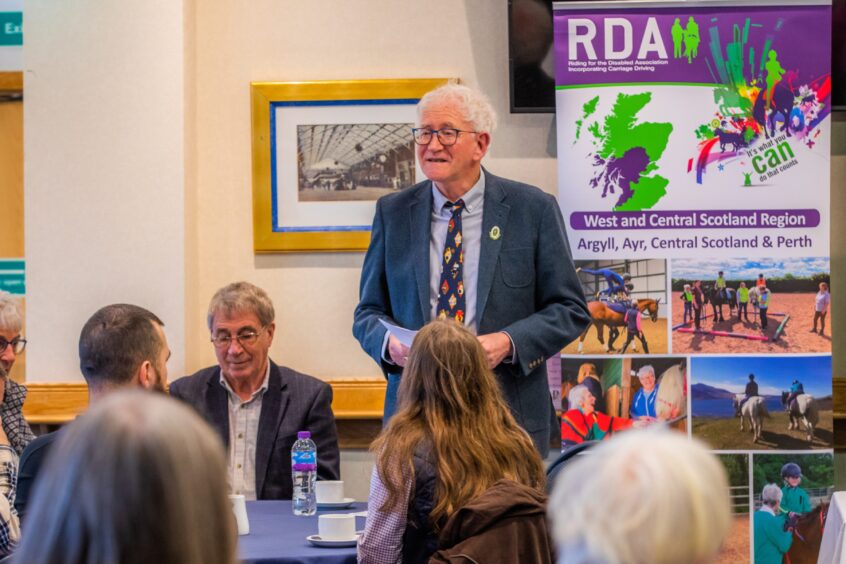 Dave Anderson in suit and tie speaking to audience in front of a Riding for the Disabled Association banner