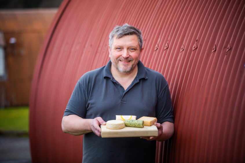 Pierre Leger smiling holding cheeseboard with cheeses next to old iron POW camp hut