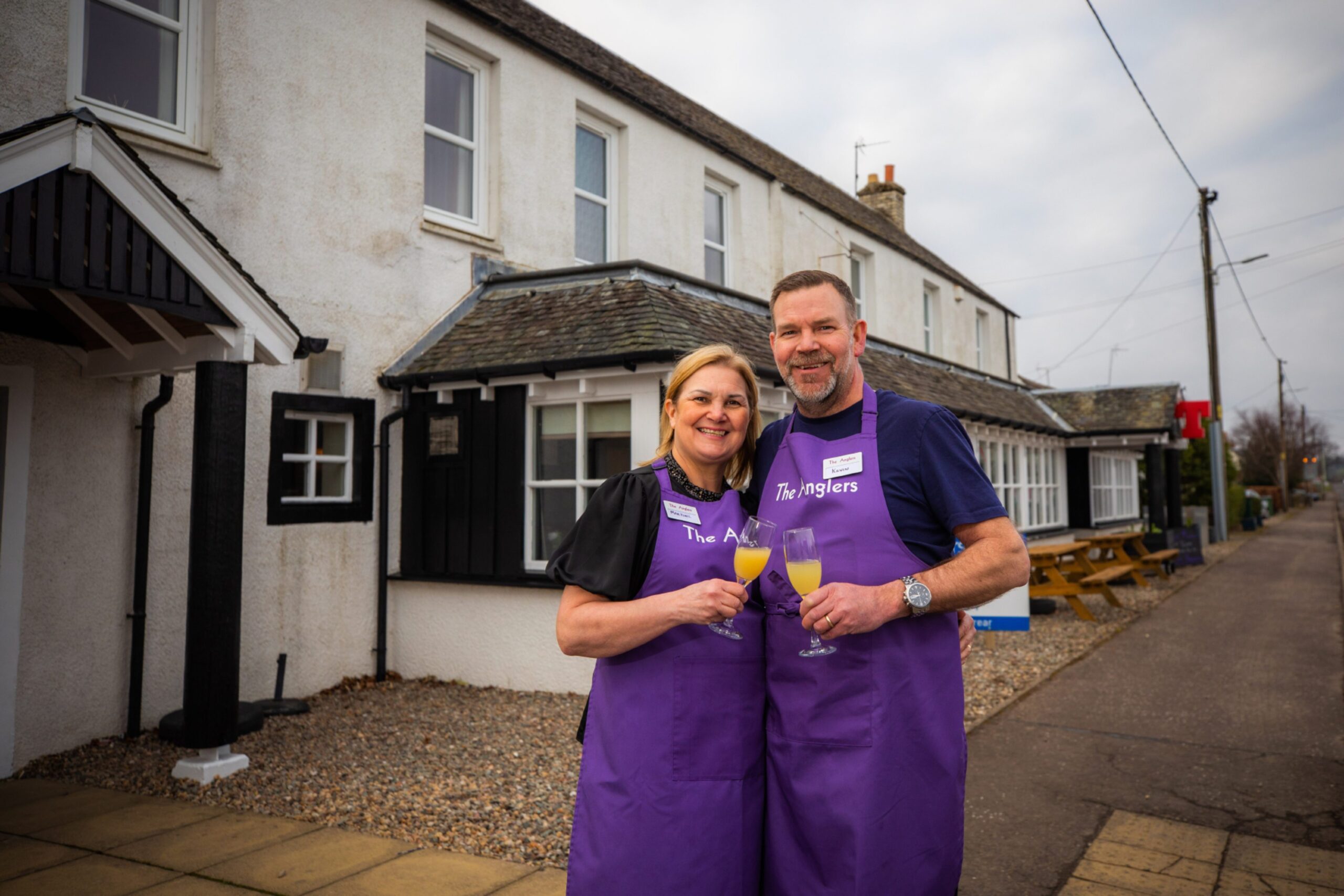 Previous tenants Martine and Kev Sinclair in purple branded aprons holding champagne flutes outside The Anglers, Guildtown