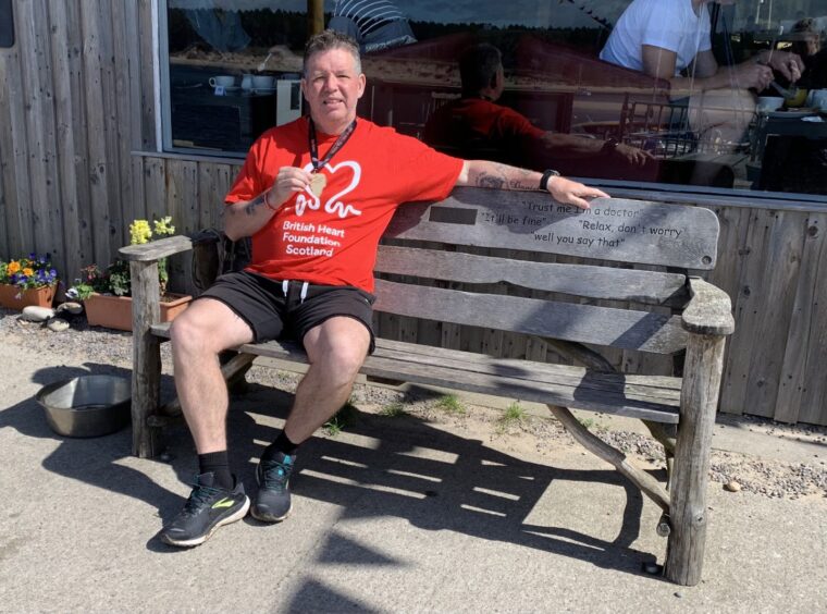 Windy WIlson in British Heart Foundation T shirt, seated on a bench holding a medal