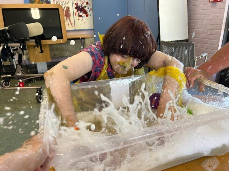 Becky enjoys water play at her local day centre, splashing her hands in a big tub of foamy water. Becky's mum say people with Angelman syndrome often love water.