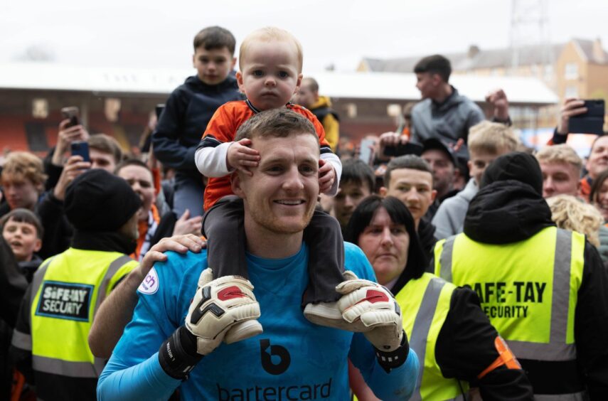 Jack Walton, with his son on his shoulders, enjoys Dundee United's title party at Tannadice, as fans crowd around him and stewards on the pitch.