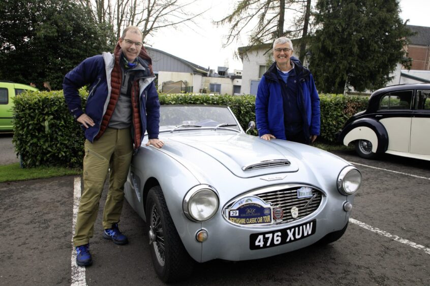 Two men standing next to small silvery blue Austin Healey car